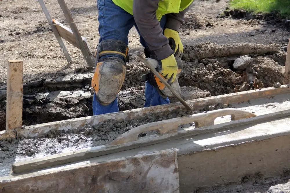 Gloved hands running a screed board along a formed edge of freshly placed concrete
